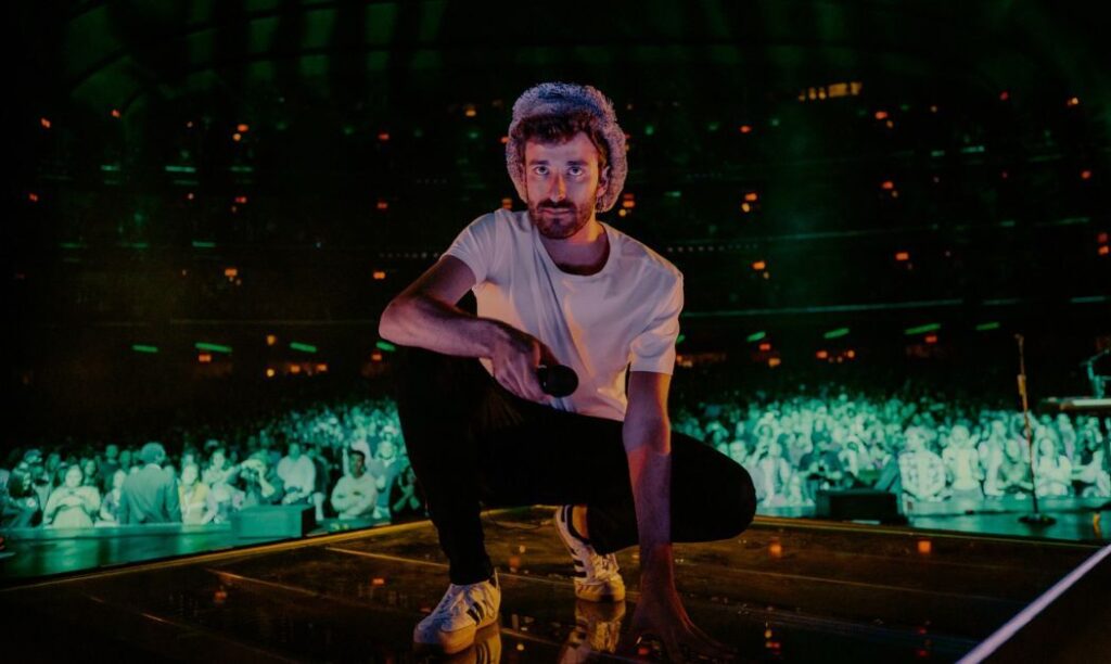 AJR band member kneeling on a stage in front of a large crowd.