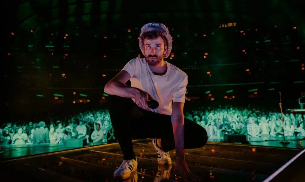 AJR band member kneeling on a stage in front of a large crowd.