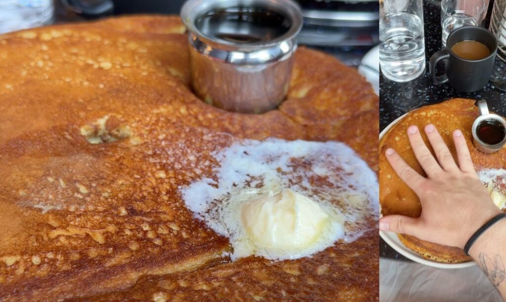 A massive golden-brown pancake at Mercury Diner in Columbus, Ohio, topped with melting butter and served with syrup; a hand is shown for scale to highlight the pancake’s enormous size